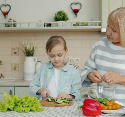 Cute little girl and elderly woman granny are cooking salad at kitchen table together preparing meal and talking. Lifestyle, nutrition and people concept.