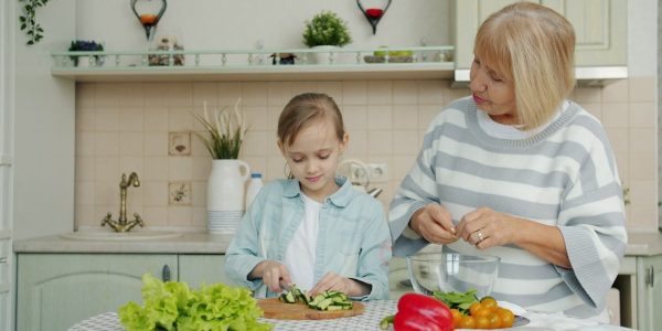 Cute little girl and elderly woman granny are cooking salad at kitchen table together preparing meal and talking. Lifestyle, nutrition and people concept.